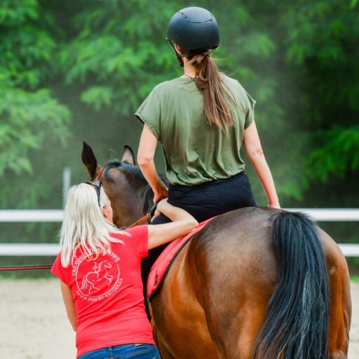 stojąca przy koniu kobieta asekurująca jeźdźca w tle padok